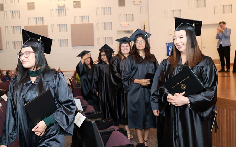 Female graduates in black caps and gowns holding diplomas and exiting auditorium